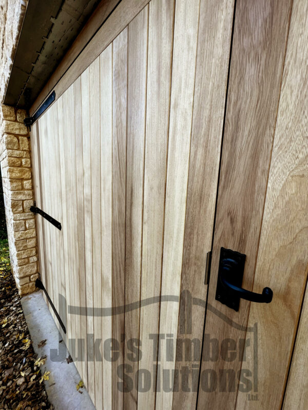 Close up of an iroko super heavy duty full board garage door, installed into a garage, with black metal handles and hinges